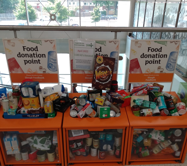 Orangene Schränke mit Lebensmittelspenden gefüllt, darunter Flaschen, Dosen und Kartons, mit Informationsschildern und Blick auf Fahrzeuge, Bäume, Gebäude und Himmel durch Glasfenster.