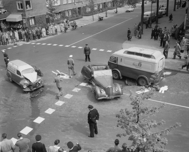 Schwarz-weißes Bild eines Autounfalls an einer Straßenkreuzung mit mehreren Fahrzeugen, einem Lieferwagen und einer Gruppe von Zuschauern, mit Gebäuden, Bäumen, Strommasten und Texttafeln im Hintergrund.