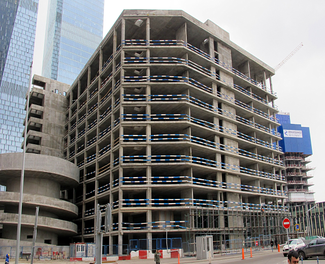 Großer Bau in einer Stadt mit Wolkenkratzern, Fahrzeugen, Fußgängern, Verkehrsinfrastruktur und Kränen unter einem klaren blauen Himmel.