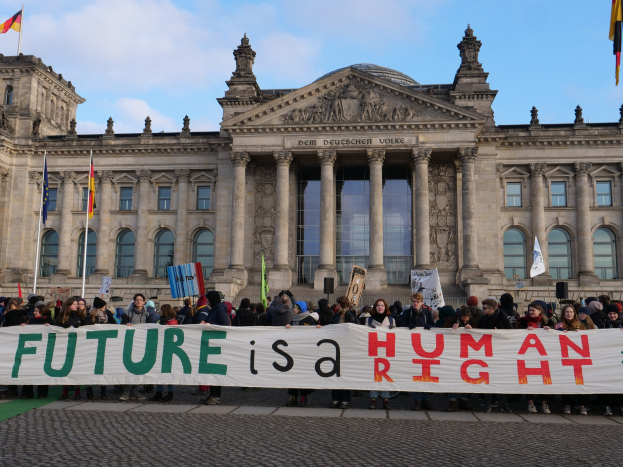 Gruppe von Menschen mit einem Transparent 'Zukunft ist ein Menschenrecht' vor dem Reichstagsgebäude in Berlin, Deutschland, mit bewölktem Himmel im Hintergrund.