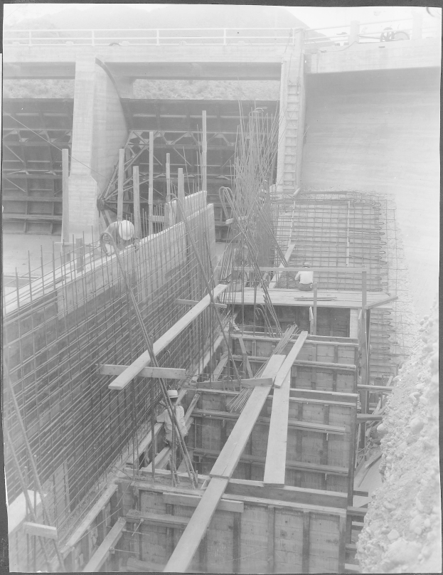 Ein Schwarz-Weiß-Foto eines Bauplatzes mit einer Brücke im Hintergrund, das Arbeiter, Stangen, hölzerne Gegenstände und andere Baustoffe zeigt, mit Text unten.