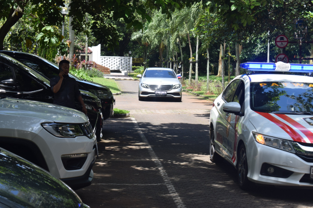 Ein Polizeiwagen steht am Straßenrand neben anderen Fahrzeugen, während eine Person in der Nähe ein Mobiltelefon hält; Bäume, Schilder, Pfosten und eine Wand sind im Hintergrund zu sehen.