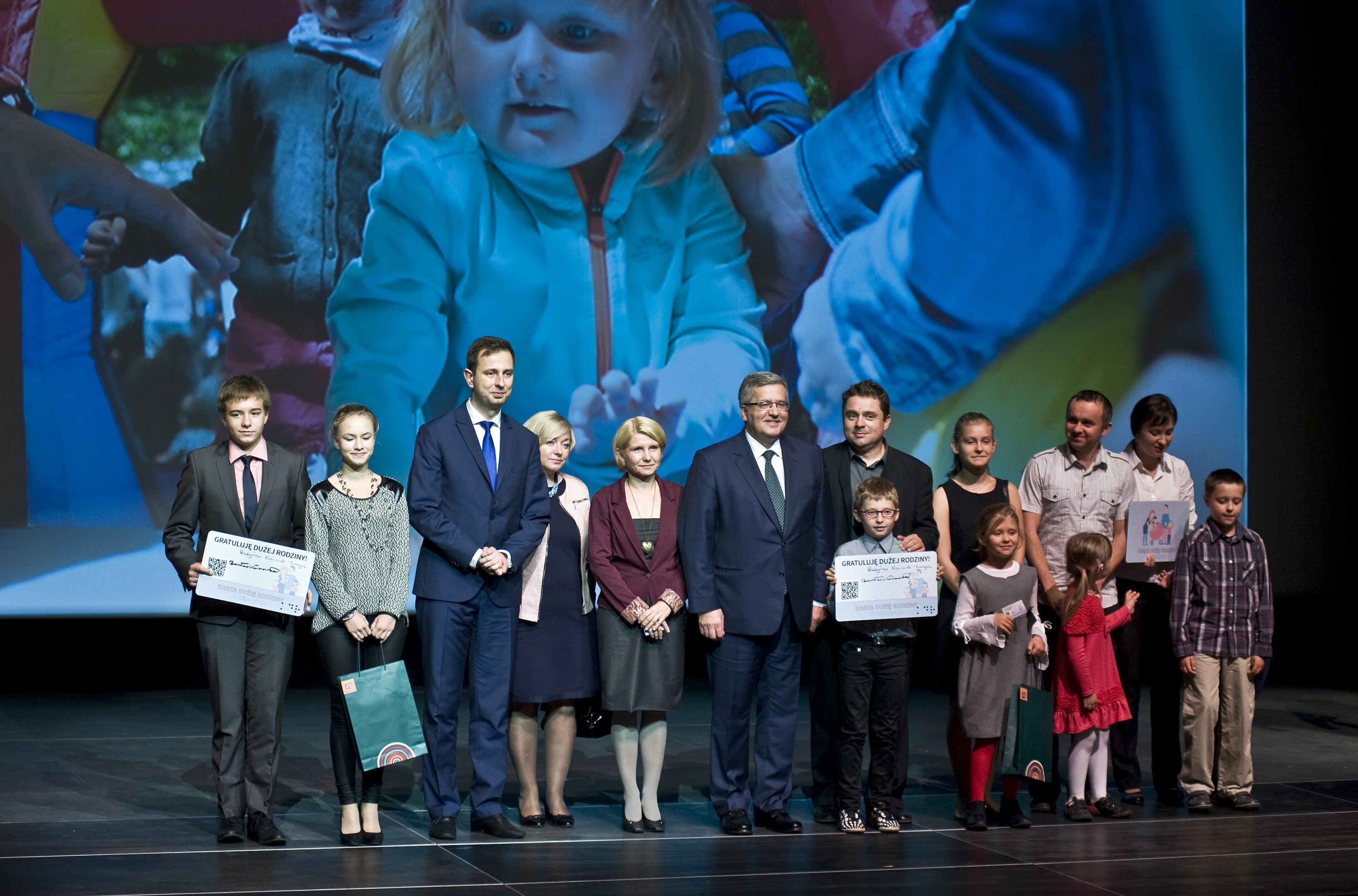 Gruppe von Menschen auf einer Bühne mit Taschen und Urkunden, im Hintergrund ein Bildschirm mit einem Kind und anderen.