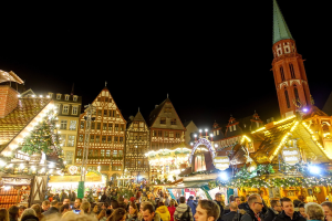 Ein geschäftiger Weihnachtsmarkt in Nürnberg, Deutschland mit Menschen um geschmützte Stände, festliche Lichter, Gebäude mit Fenstern, einen Uhrturm und einen dunklen Himmel.