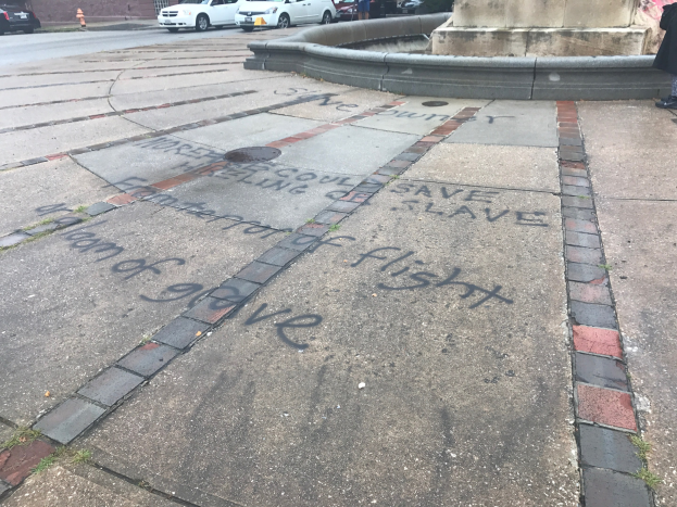 Gesteig mit 'Black Lives Matter'-Graffiti vor einem Gebäude, umgeben von Fahrzeugen, Menschen, Bäumen, Pfählen und einer Wand.