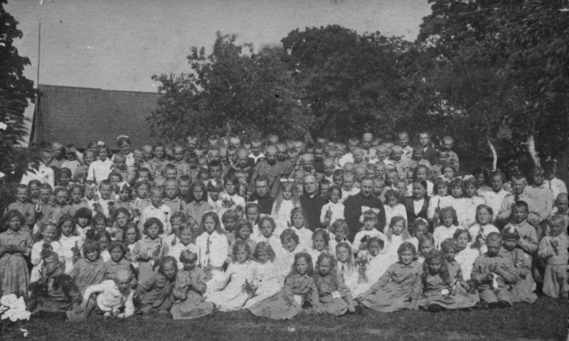 Schwarzes und weißes Foto einer Gruppe von Kindern, die vor einem Haus mit Bäumen und einem klaren Himmel posieren.