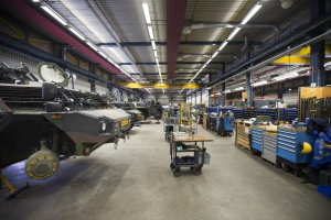 Großlager mit Maschinen, Fahrzeugen auf der linken Seite, Transportwagen mit Gegenständen, Holzboxen und Deckenbeleuchtung.