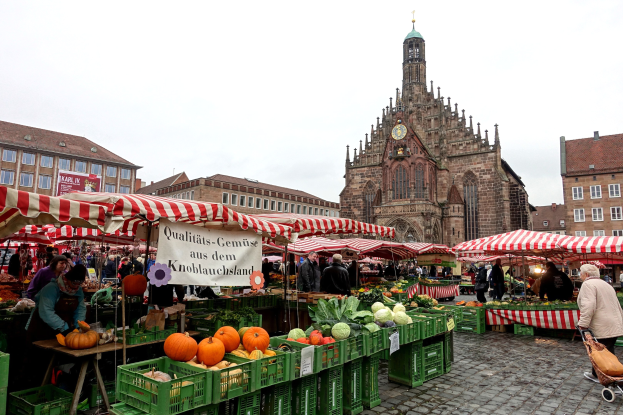 Ein belebter Markt in Nürnberg, Deutschland, mit Ständen, die verschiedene Früchte und Gemüse anbieten, Menschen, die herumlaufen, und Zelten, mit Gebäuden und einem Uhrenturm im Hintergrund unter einem sichtbaren Himmel.