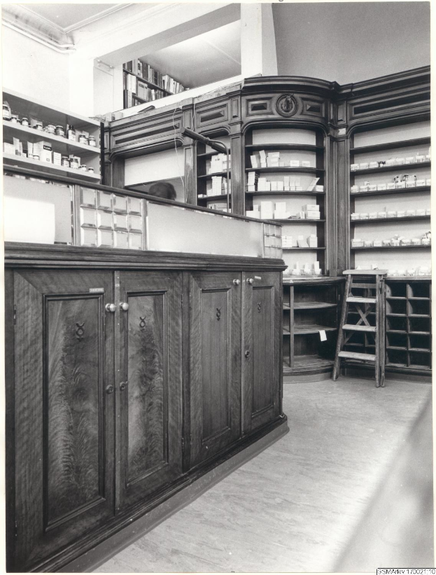 Schwarz-weiß-Foto eines vintage-apothekeninneren mit Theke, Schränken, einer Leiter, Regalen und Text oben.