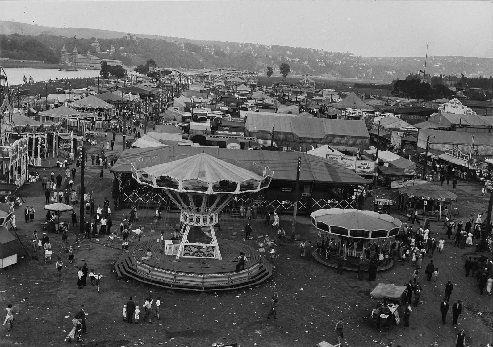 Ein altes Schwarz-Weiß-Foto eines pulsierenden Volksfestes mit einem Karussell in der Mitte, umgeben von Menschen, Buden, Pfählen, Bäumen, Gebäuden und einer Brücke im Hintergrund unter einem Himmel.