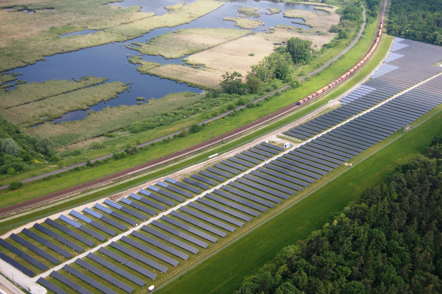 Luftaufnahme eines Solarparks mit Panelen in einer grünen Wiese, umgeben von Bäumen und Wasser, mit einem Zug auf einer nahen Schiene.