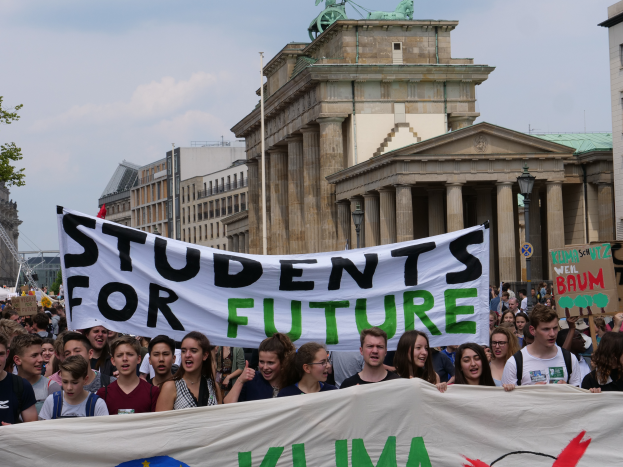 Eine Gruppe von Schülern marschiert in Berlin, die ein buntes Banner mit der Aufschrift 'Students for Future' tragen, mit Gebäuden, Bäumen und Himmel im Hintergrund.