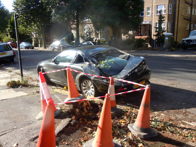 Verunfalltes Auto am Stra√enrand mit orangen Verkehrskegeln, Bäumen, Gebäuden und einem klaren blauen Himmel im Hintergrund.