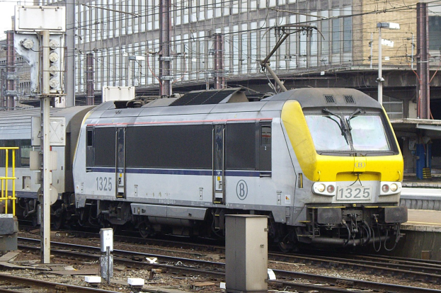 Diesellokomotive mit hydraulischem Antrieb fährt auf Schienen neben einem hohen Gebäude, mit Strommasten und -drähten, einer Plattform auf der rechten Seite und einem klaren blauen Himmel.