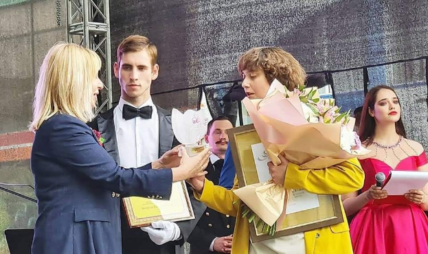 Gruppe von Menschen auf einer Bühne, eine in gelbem Kleid mit Blumen, eine andere mit Papieren und Mikrofon, links ein Mann im blauen Blazer und weißem Hemd, der einen Preis erhält.