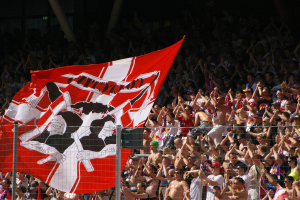 Große Menschenmenge in einem Stadion mit einer roten und weißen Flagge, Maschendrahtzaun im Vordergrund und Säulen vor einem dunklen Hintergrund.