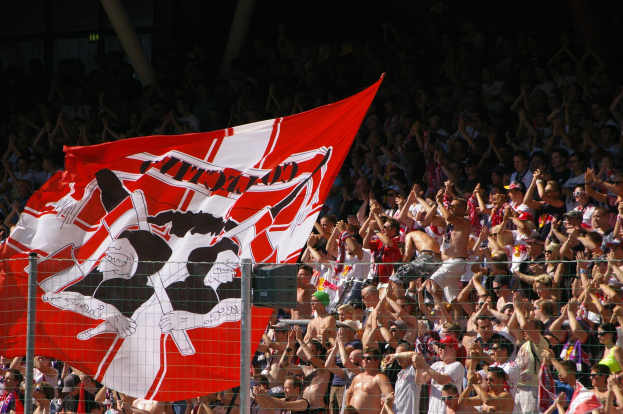 Große Menschenmenge in einem Stadion mit einer roten und weißen Flagge, Maschendrahtzaun im Vordergrund und Säulen vor einem dunklen Hintergrund.