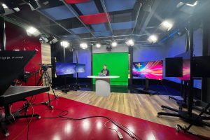 Ein Mann sitzt an einem Schreibtisch in einem TV-Studio mit einer grünen Wand, einem Laptop und anderen Gegenständen, mit Kameras und Equipment links und Bildschirmen und Deckenlampen im Hintergrund.