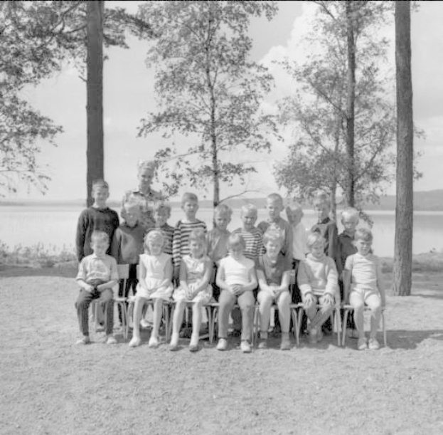 Schwarz-weiß-Foto einer Gruppe von Kindern, die auf Stühlen sitzen und vor einem See stehen, mit Bäumen, Pflanzen, Hügeln und einem bewölkten Himmel im Hintergrund und Text unten, der "1940er-Kinder am See" lautet