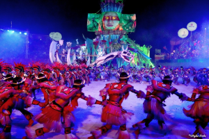 Eine Gruppe von Menschen in bunten Kostümen tanzt auf einer Bühne während des Rio-Karnevals 2014, mit einer Menge, die auf Stühlen sitzt, beleuchtet von Lichtern und einem Bildschirm, der eine Person und andere Gegenstände anzeigt.