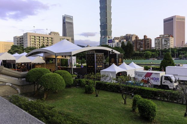 Eine Gruppe von Zelten in einem Park mit Grünflächen, Menschen, Fahrzeugen, Gebäuden und einem klaren Himmel, wahrscheinlich während des Hong Kong International Film Festivals.