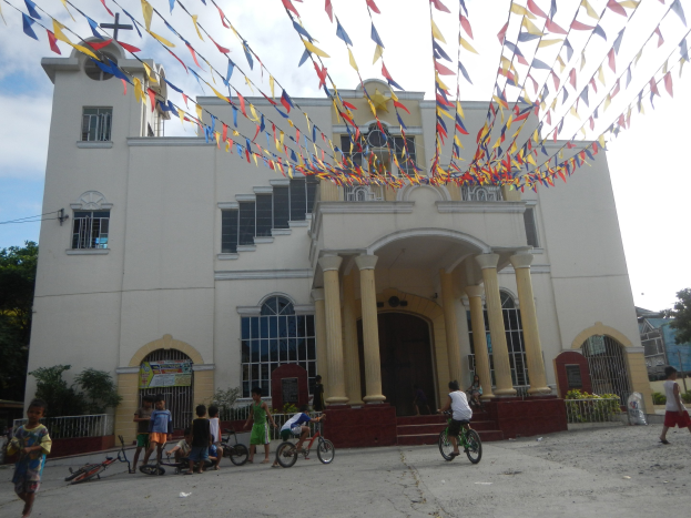 Eine Gruppe von Menschen, die Fahrräder vor einer weißen Kirche mit architektonischen Details wie Fenstern, Säulen, Bögen, Türen und einem Kreuzsymbol fährt, mit Pflanzen, einem Zaun, einer Fahne, Bäumen und einem bewölkten Himmel im Hintergrund und bunten Fahnen, die an einem Seil über der Kirche befestigt sind.