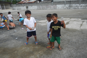 Gruppe junger Jungs, die in einer Parklötte stehen, einige halten Flaschen, mit Häusern, Bäumen und Mästen im Hintergrund unter einem klaren blauen Himmel.