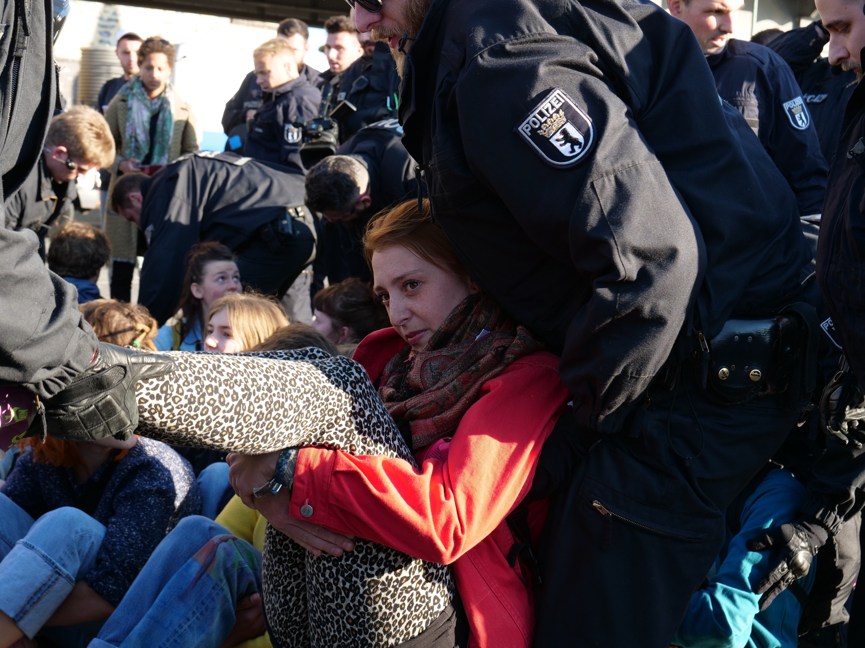 Eine Frau in dunkler Kleidung wird von einem Polizeibeamten in Uniform begleitet, hinter ihnen eine Gruppe sitzender und stehender Menschen, ein Schuppen und ein klarer blauer Himmel im Hintergrund.