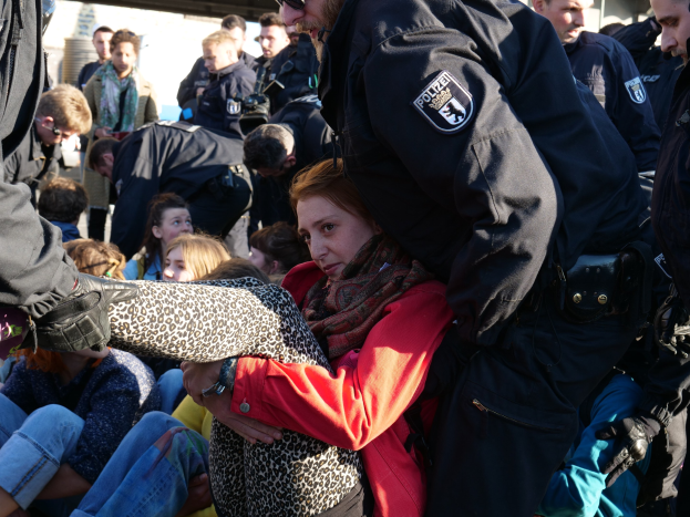 Eine Frau in dunkler Kleidung wird von einem Polizeibeamten in Uniform begleitet, hinter ihnen eine Gruppe sitzender und stehender Menschen, ein Schuppen und ein klarer blauer Himmel im Hintergrund.