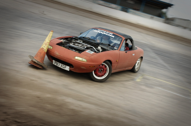 Eine Person fährt ein rotes Auto auf einer Rennstrecke mit hoher Geschwindigkeit und einem Verkehrskegel vor sich.