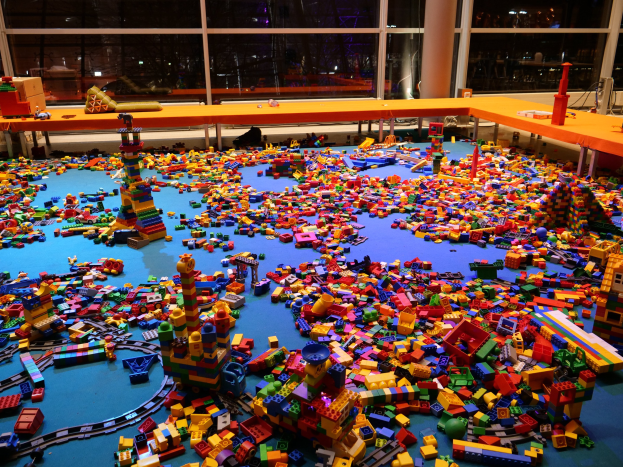 Ein großer Raum mit bunten LEGO-Bausteinen, die auf dem Boden verstreut sind, umgeben von Tischen und Säulen, mit Glasfenstern im Hintergrund, die Menschen und Lichter zeigen.