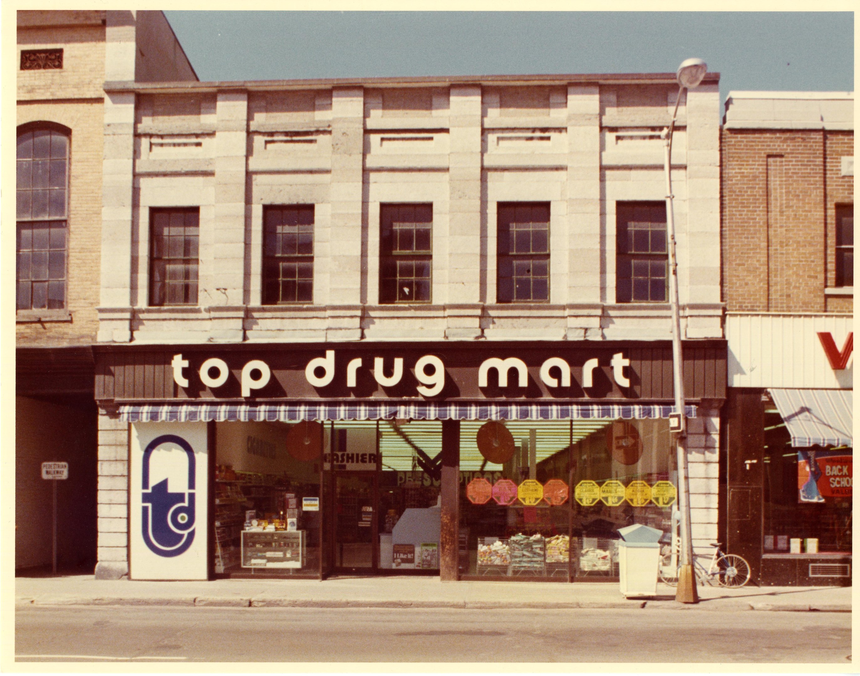 Straßenecke mit einem "Top Drug Mart"-Ladenlokal, Fenstern, einer Schautafel, einem Straßenpfosten, einem abgestellten Fahrrad, einem Müllcontainer und einem bewölkten Himmel.