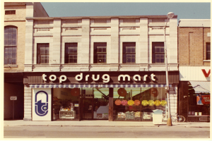 Straßenecke mit einem "Top Drug Mart"-Ladenlokal, Fenstern, einer Schautafel, einem Straßenpfosten, einem abgestellten Fahrrad, einem Müllcontainer und einem bewölkten Himmel.