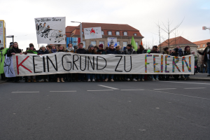 Eine Gruppe von Menschen, die auf einer Straße stehen und ein Transparent mit der Aufschrift "Kein Grund zu Feiern" halten, vor dem Hintergrund von Gebäuden, Laternenmasten, Bäumen und einem klaren blauen Himmel.