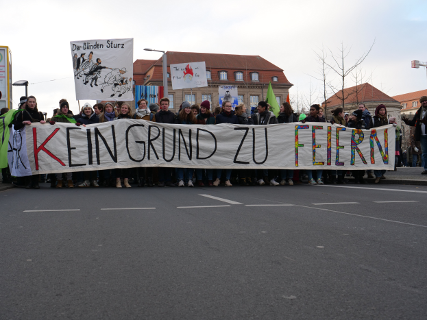 Eine Gruppe von Menschen, die auf einer Straße stehen und ein Transparent mit der Aufschrift "Kein Grund zu Feiern" halten, vor dem Hintergrund von Gebäuden, Laternenmasten, Bäumen und einem klaren blauen Himmel.
