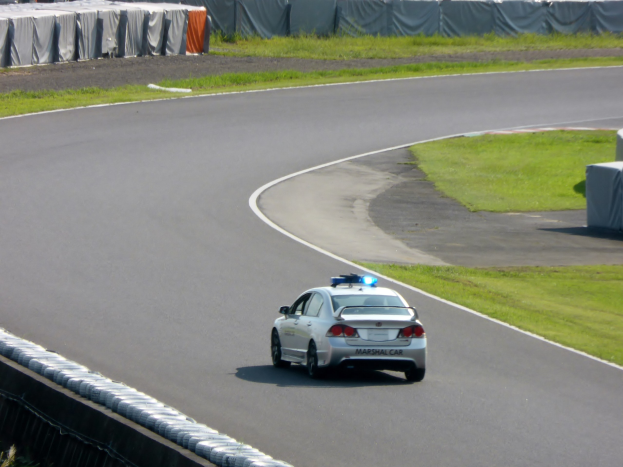 Polizeiauto auf einer Rennstrecke mit Gras auf beiden Seiten, mit Zelten und Menschen im Hintergrund.