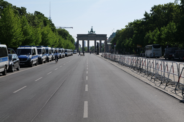 Lange Reihe von Polizeiwagen auf der Straße vor dem Brandenburger Tor in Berlin, Deutschland geparkt, mit Menschen auf Fahrrädern und Barrieren auf der Straße, Bäume an den Seiten und ein Tor mit Statuen im Hintergrund.