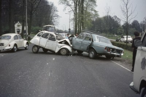 Zwei beschädigte Autos am Straßenrand mit Menschen in der Nähe, umgeben von Bäumen, Pfählen, Gebäuden und einem klaren blauen Himmel.