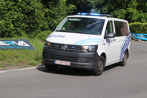 Ein Polizeiwagen mit weißer, blauer und schwarzer Lackierung fährt auf einer Straße neben einem Wald, mit Gras und einem Schild am Straßenrand und Bäumen im Hintergrund.