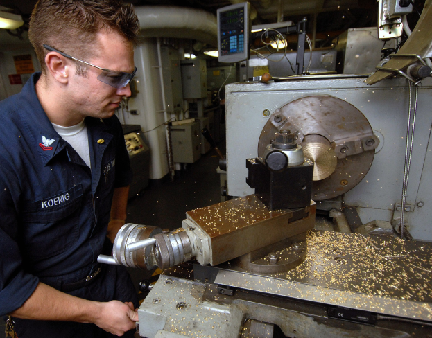 Ein Monteur mit Brille bedient eine Maschine in einer Fabrik mit sichtbaren Lampen, Rohren, Maschinen und einem Bildschirm im Hintergrund.