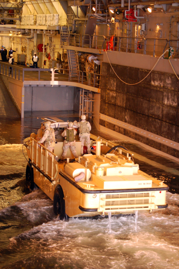 Amphibisches Fahrzeug mit Helmträgern, die durch Wasser fahren, neben einer Wand, Geländern, Lampen und Rohren im Hintergrund.