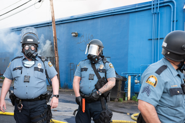 Drei Polizisten in Einsatzausrüstung, darunter Helme und Gasmasken, gehen eine Straße mit einer blauen Wand, Rohren und einem Strommast mit Drähten im Hintergrund entlang, unter einem sichtbaren Himmel.