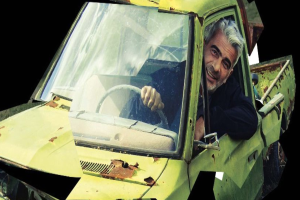 Ein Mann lächelt hinter dem Steuer eines gelben, rostigen und verbeulten Autos