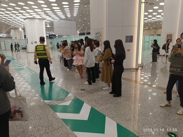 Menschen in der Schlange an einem Flughafen-Sicherheits-Check-in-Bereich, einige mit Taschen und Mobiltelefonen, mit Säulen und Deckenleuchten im Hintergrund.