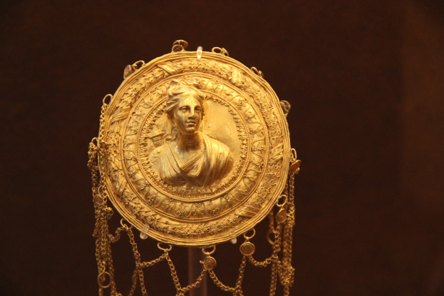 Goldene Medaille mit einer Frauengesicht, das detaillierte Verzierungen zeigt, vor einem dunklen Hintergrund.