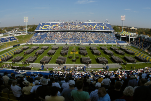 Eine große Menschenmenge in einem Stadion, die ein Fußballspiel schaut, wobei einige stehen und andere sitzen, auf einem grünen Rasen unter einem klaren blauen Himmel, umgeben von Laternenmasten, Bäumen und Gebäuden.