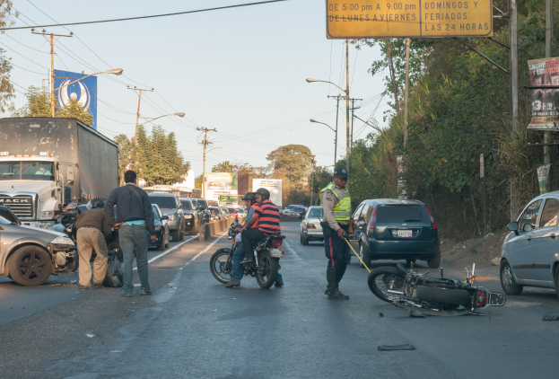 Eine Gruppe von Menschen steht um ein verunglücktes Motorrad auf der Seite einer Straße mit mehreren Fahrzeugen, Bäumen, Pfählen, Laternen, Schildern und dem Himmel im Hintergrund.