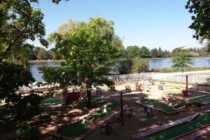 Minigolf-Platz in einem Park mit grünem Gras, Bäumen und einem Zaun, umgeben von verstreuten Stühlen und Tischen, mit einem Gewässer und einem Boot im Hintergrund unter dem Himmel.