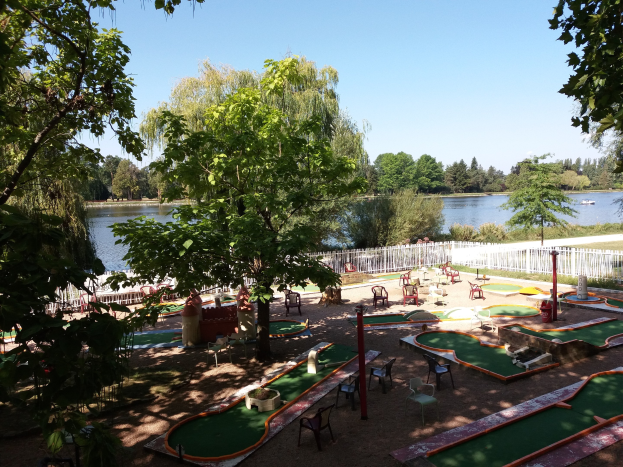 Minigolf-Platz in einem Park mit grünem Gras, Bäumen und einem Zaun, umgeben von verstreuten Stühlen und Tischen, mit einem Gewässer und einem Boot im Hintergrund unter dem Himmel.
