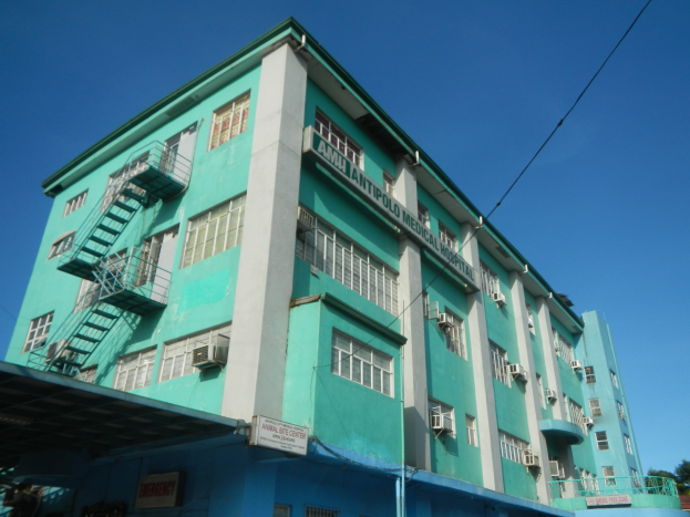 Ein großes grünes Gebäude mit vielen Fenstern, Klimaanlagen und Treppen, umgeben von Bäumen, Fahrzeugen und Strommasten unter einem klaren blauen Himmel.